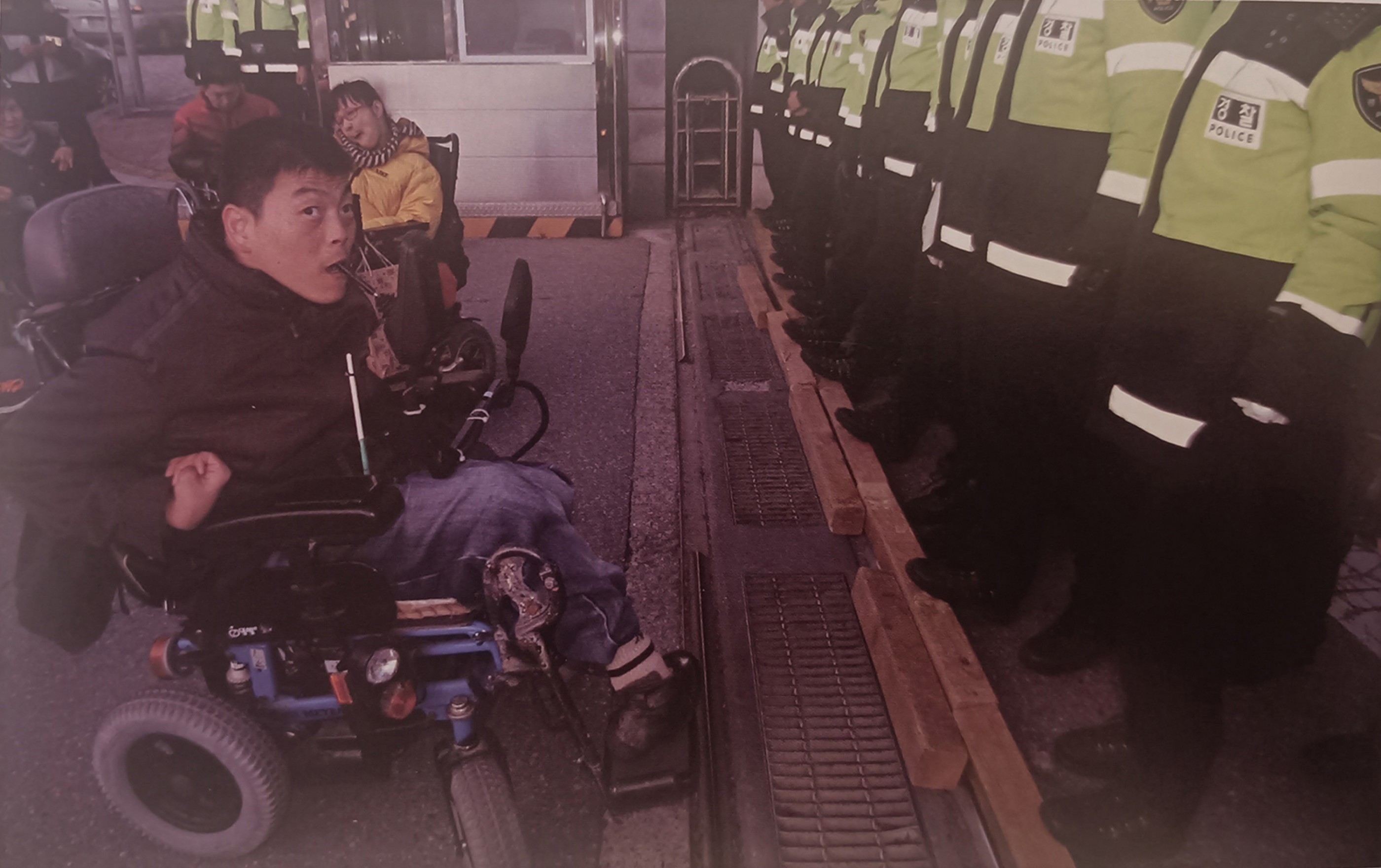 <p>Police block a wheelchair with wooden sticks. Scan from Photo Album <em>&quot;Leave No One Behind: 10-year Record of Solidarity Against Disability Discrimination&quot;</em> 2007-2022.</p>